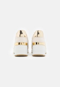 Baskets beiges avec un accent doré brillant au talon, semelle blanche et tissu texturé. Forme arrondie avec une finition lisse.