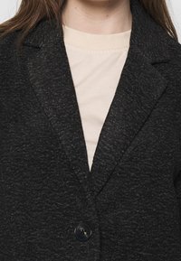 Skräddarsydd svart kavaj tillverkad av texturerad tyg, med slagkrage och en ensam mörk knapp, bärs över en ljusbeige skjorta.