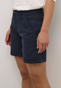 Tumesinised puuvillased lühikesed püksid, millel on ees taskud, keeratud äär ja nööbiga kinnitamine pihtjal, et tagada kindel istuvus.