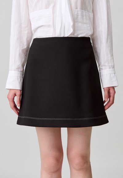 Gonna nera a linea ad A con una texture liscia, orlo dritto e cuciture in contrasto bianche. Abbinata a una camicetta bianca a maniche lunghe.