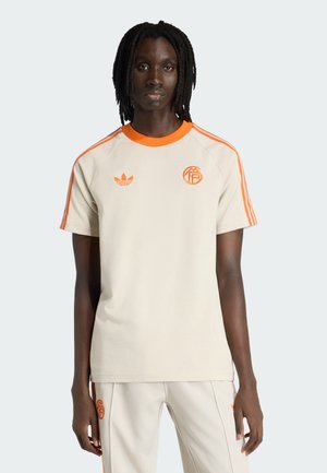 Person mit dunkler Hautfarbe und geflochtenem Haar, bekleidet mit beiger und oranger Adidas-Sportbekleidung, stehend vor einem schlichten hellen Hintergrund.