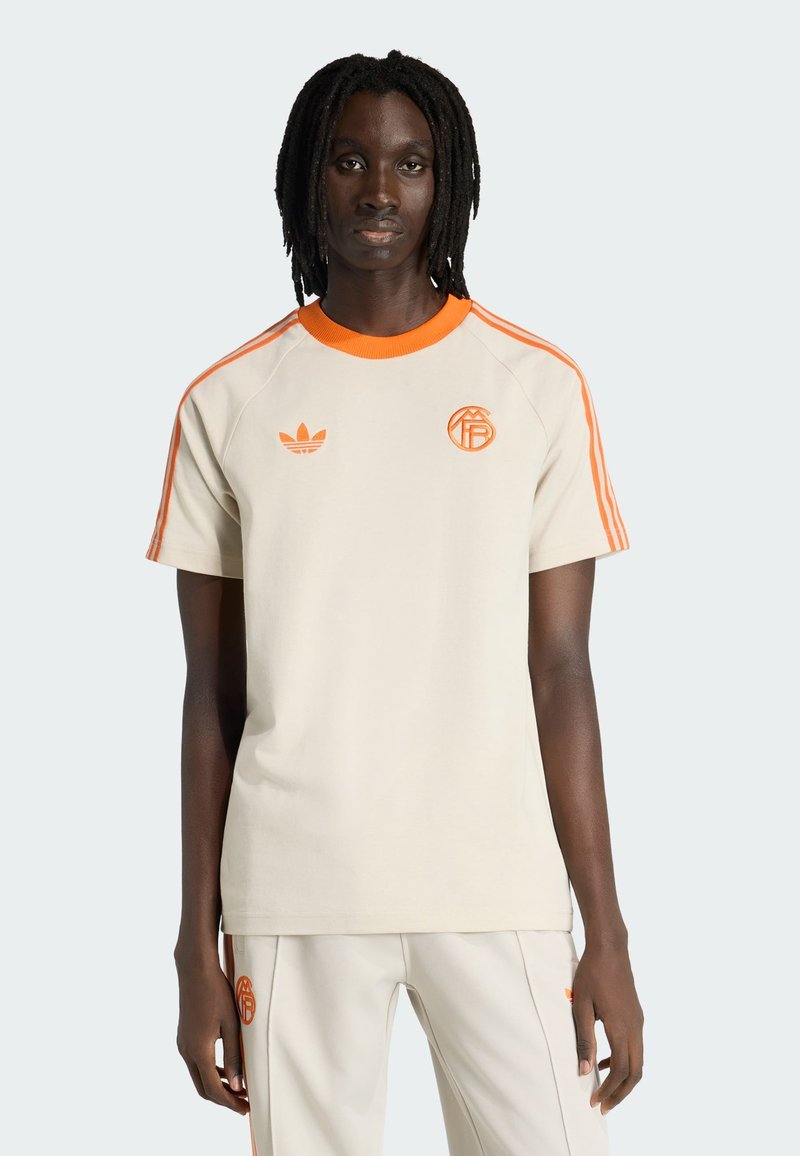 Άτομο με σκουρόχρωμο δέρμα και κοτσίδες, ντυμένο με αθλητικά ρούχα Adidas σε μπεζ και πορτοκαλί χρώμα, στέκεται μπροστά σε απλό φωτεινό φόντο.