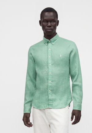 Homme portant une chemise vert clair à manches longues boutonnée et un pantalon blanc, debout devant un fond clair uni.