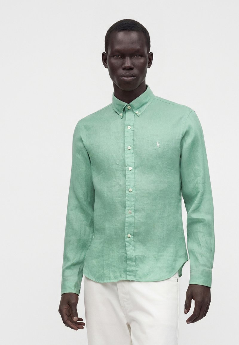 Homme portant une chemise vert clair à manches longues boutonnée et un pantalon blanc, debout devant un fond clair uni.