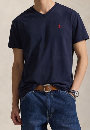 Man draagt een donkerblauw V-hals T-shirt met rood geborduurd logo, bruine riem, blauwe spijkerbroek en polshorloge met logo bandje, handen in de zakken.