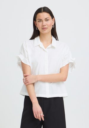 Ung kvinde med langt mørkt hår iført en hvid kortærmet bluse med flæsemanchetter og sorte bukser, stående med armene over kors.