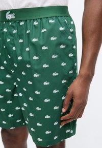 Shorts verts avec logos de crocodile blancs, taille élastique et poches latérales, portés par une personne à la peau foncée et portant une chemise blanche.