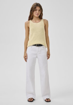 Mujer de pie con top de punto sin mangas beige, pantalones blancos de pierna ancha, cinturón negro y chanclas negras sobre fondo liso.