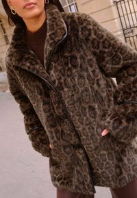 Leopárdmintás műszőrme kabát sálgallérral, puha tapintással, fekete kiegészítőkkel és gombos záródásokkal. Barna és bézs színösszeállítás.