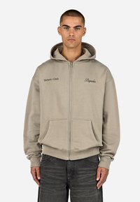 Felpa beige con zip e tasca frontale, con la scritta "Drivers Club" e "Pegador" ricamate in nero. Tessuto morbido, vestibilità comoda.