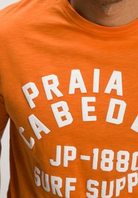 Orange shirt with white block letters spelling "PRAIA CABEDELA JP-1880 SURF SUPP" on the chest, worn by a bearded person.