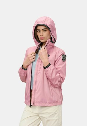 Donna che indossa una giacca con cappuccio rosa chiaro con una toppa con logo sulla manica, che la chiude con la zip, vestita con pantaloni chiari e una camicia blu.