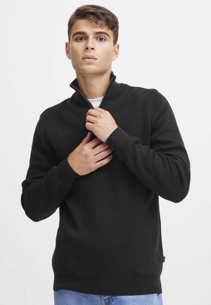 Schwarzer Strickpullover mit einem Reißverschlusskragen, gerippten Bündchen und Saum. Das Model hält den Kragen und trägt ein weißes T-Shirt darunter, kombiniert mit blauen Jeans.