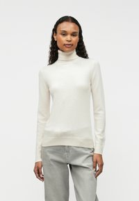 Cream turtleneck sweater in soft, fine knit material with ribbed cuffs and hem, paired with light gray trousers.