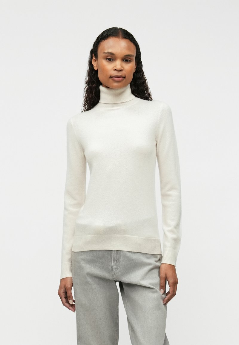 Cream turtleneck sweater in soft, fine knit material with ribbed cuffs and hem, paired with light gray trousers.