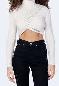 Maglione bianco a coste, cropped, con collo alto e design a incrocio diagonale, abbinato a jeans neri a vita alta con dettagli a pulsante laterali.