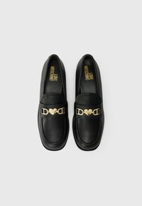 Mocassins em pele preta com um detalhe em hardware em forma de coração dourado e uma textura suave, apresentando a marca nas palmilhas.