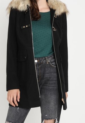 Woman wearing a black coat with fur collar over a teal textured sweater and ripped faded black jeans, standing against a plain background.