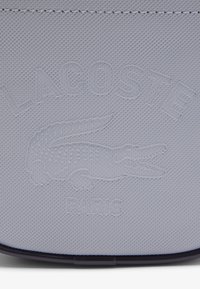 Pochette grigia chiara con texture, caratterizzata dal logo in rilievo "LACOSTE" e dal coccodrillo, con "PARIS" sottostante. Materiale liscio e dettaglio di finitura scura.