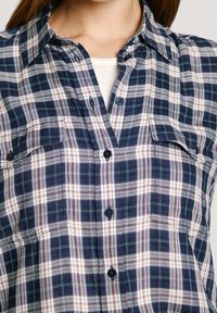 Kvinna som bär en skjorta i marinblå och vit rutmönster över en vit topp, som visar kragen och bröstområdet.