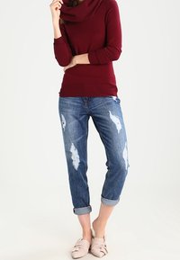 Maglione color granata con collo a cappuccio abbinato a jeans blu strappati. I jeans presentano strappi e risvolti arrotolati. Scarpe piatte rosa chiaro completano l'outfit.