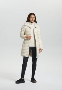 Lichtbeige pufferjas met horizontale stiknaden, capuchon en voorkant rits. Gecombineerd met zwarte leggings en schoenen. Glad oppervlak en middellange uitvoering.