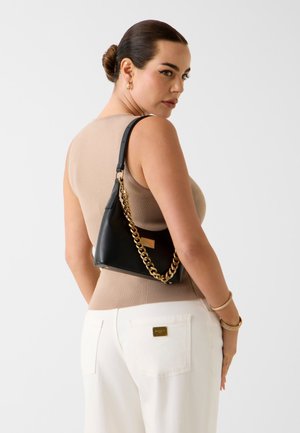 Donna con capelli scuri raccolti in uno chignon, indossa una canottiera beige, pantaloni bianchi, gioielli d'oro e tiene una borsa nera con tracolla a catena d'oro sulla spalla.