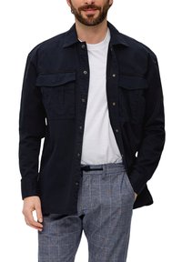 Camicia blu navy con bottoni in cotone, dotata di due tasche sul petto e una vestibilità rilassata, indossata sopra una maglietta bianca e pantaloni a quadri.
