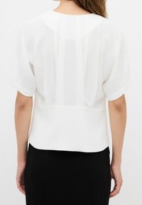 Femme portant un chemisier blanc ample à manches courtes avec des plis dans le dos, associé à un pantalon noir taille haute.