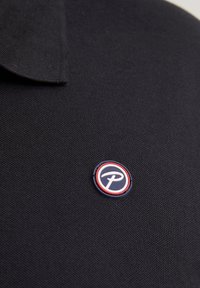 Mustapallo paita, jossa on teksturoitu kangas. Vasemmassa rinnassa on pyöreä logo punaisessa, valkoisessa ja sinisessä värissä. Lyhyt kaulus.