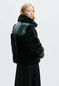 Ciemnozielona ocieplana kurtka z błyszczącym czarnym panelem na ramieniu, wysokim kołnierzem i puszystą teksturą. Nosi ją osoba w długiej czarnej spódnicy.