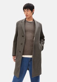 Grau melierter Woll-Mix-Mantel mit taillierter Passform, drei Knöpfen, Reverskragen, Seitentaschen und klassischer Silhouette. Übereinander getragen über einem braunen Pullover.
