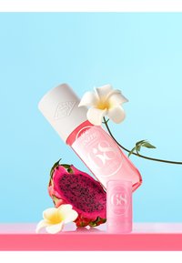 Różowy spray perfumowany i balsam Sol de Janeiro Cheirosa 68 z białymi kwiatami plumeryi i plasterkami owocu smoka na różowej powierzchni.