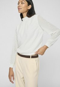 Blusa bianca con texture e motivo a zigzag, colletto alto e polsini elasticizzati, abbinata a pantaloni beige e una cintura marrone scuro.