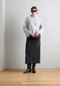 Šedý hrubý pletený svetr s rolákem, zkombinovaný s dlouhou černou džínovou sukní a černými kozačkami po kolena. Modelka drží kulatou černou tašku.