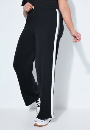 Person wearing black wide-leg pants with a white side stripe and white sneakers, standing against a white background.