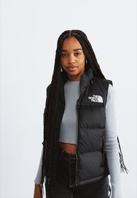 Fată tânără cu cosițe lungi, purtând o vestă puffer neagră peste un top scurt gri și pantaloni negri, stând în fața unui fundal deschis simplu.