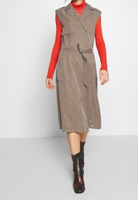 Khakifarbene ärmellose Kleid mit einem gegürteten Taillenbereich, seitlichen Taschen und einer lockeren Passform, kombiniert mit schwarz-roten gemusterten Stiefeln. Darunter ein rotes Langarmoberteil.