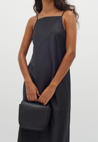 Czarna, bezrękawnikowa sukienka z prostym dekoltem. Trzyma czarną skórzaną torbę na ramię z klapką i zaokrąglonymi rogami.