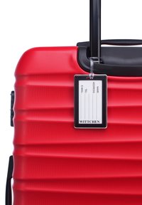 Valise rouge à roulettes avec une surface texturée, poignée noire et une étiquette de bagages attachée, comportant un emplacement pour les informations personnelles.