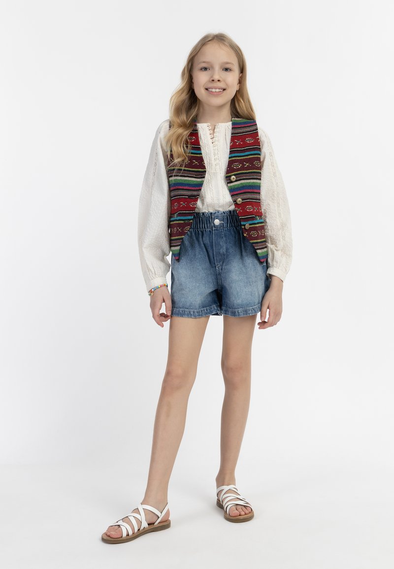 Mädchen trägt eine weiße Bluse, eine bestickte rot- und mehrfarbige Weste, Jeansshorts und weiße Sandalen mit Riemen. Glatter Stoff und lässiger Stil.