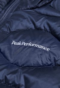 Tamno plava puffer jakna s kvadratnim uzorkom, s "PeakPerformance" logotipom u bijeloj izvezenoj boji na prednjoj strani. Vodootporan materijal.
