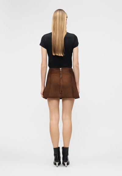 Γυναίκα με ίσια ξανθά μαλλιά στέκεται με την πλάτη, φοράει μαύρο τοπ με κοντά μανίκια, καφέ σουέντ μίνι φούστα με φερμουάρ στο πίσω μέρος και μαύρα μποτάκια μέχρι τον αστράγαλο.