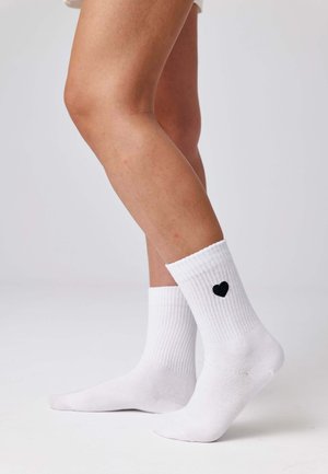 Jambes portant des chaussettes blanches côtelées avec un petit cœur noir sur le côté extérieur, debout devant un fond clair uni.