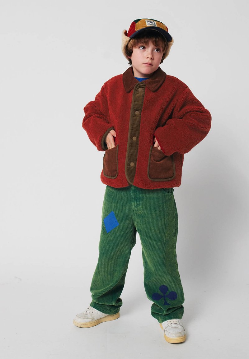 Giacca rossa pelosa con accenti in corduroy marrone, pantaloni in corduroy verdi con toppe blu e un berretto colorato con alette per le orecchie. Scarpe da ginnastica bianche.