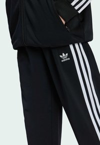 Černý teplákový komplet se bílými pruhy podél rukávů a stran kalhot, s logem Adidas na levé nohavici. Vyrobeno z hladké tkaniny.