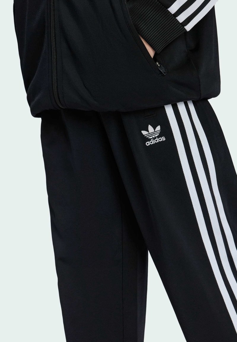Černý teplákový komplet se bílými pruhy podél rukávů a stran kalhot, s logem Adidas na levé nohavici. Vyrobeno z hladké tkaniny.