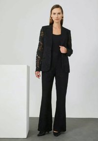 Black lace blazer with structured design, layered over a black top and flared trousers. Sophisticated texture and tailored fit.
