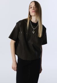 Una donna con capelli biondi lisci indossa un top in pelle nera largo con dettagli in borchie e una collana con un grande pendente a forma di cuore in argento.