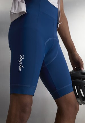 Κοντινό πλάνο ενός ατόμου που φοράει μπλε ποδηλατικό σορτς με το λογότυπο "Rapha", κρατώντας ένα μαύρο κράνος ποδηλάτου στο πλάι.